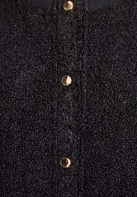 Melna teksturēta auduma gabals ar trim spīdīgām zelta pogām, kas vertikāli izkārtotas centrā.