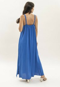 Robe maxi bleue avec des fines bretelles, en tissu fluide et avec un dos froncé. Elle présente une coupe longue et ample, avec des fentes sur les côtés et une texture lisse.