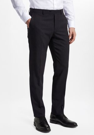 Pantalon formel noir porté avec des chaussures habillées noires et une chemise blanche à manches longues, mains détendues le long du corps sur fond blanc.
