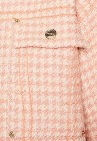 Růžový a bílý tweedový kabát s houndstooth vzorem, s odznaky zlatého kovu a oranžovými švy.