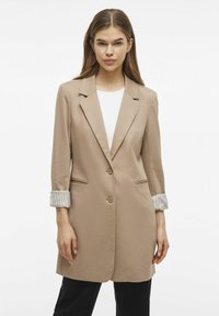 Blazer beige su misura con un solo bottone, rever a punte e maniche lunghe a righe arrotolate. Presenta una texture morbida e tasche laterali.