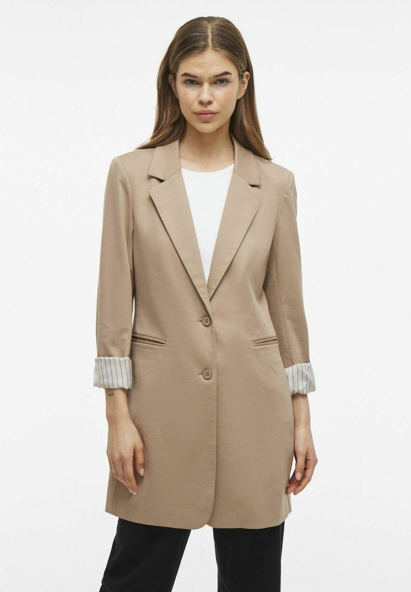 Blazer beige su misura con un solo bottone, rever a punte e maniche lunghe a righe arrotolate. Presenta una texture morbida e tasche laterali.