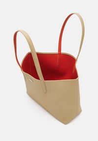 Geantă tote maronie cu un exterior texturat, interior roșu și mânere lungi, plate. Prezintă un logo mic pe o parte. Design deschis, fără închidere.