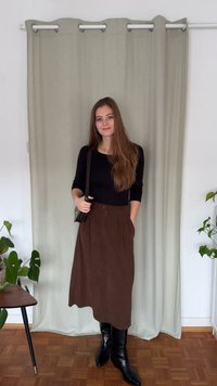 Jupe longue marron avec des plis et des poches, associée à un haut noir ajusté et des bottes noires montantes jusqu'au genou. En arrière-plan, un rideau vert clair.