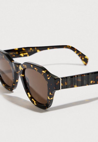 Hnědé sluneční brýle v želvím stylu s kulatým tvarem, silným acetátovým rámem a pruhovanými zdobenými nožičkami. Čočky jsou tmavě hnědé.