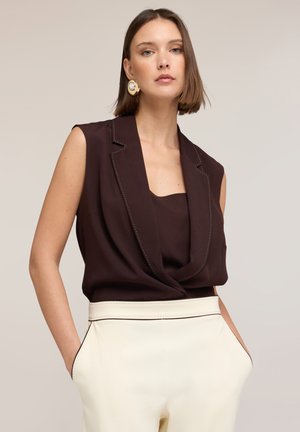 Kvinde med kort brunt hår iført ærmeløs mørkebrun blazer-lignende top og højtaljede off-white bukser, hænderne i lommerne, neutralt baggrund.