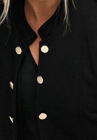 Czarna kurtka z wysokim kołnierzem, z złotymi guzikami. Miękka tkanina o subtelnej fakturze, podkreślająca dopasowany krój wokół dekoltu.