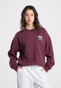 Sweat-shirt oversize bordeaux avec encolure côtelée, manches longues et un petit patch graphique. Associé à un pantalon de survêtement gris clair. Texture douce.
