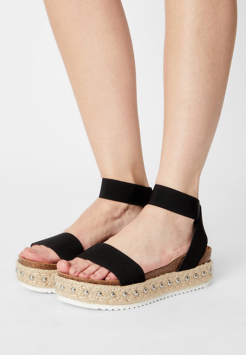 Sandales espadrilles noires avec des sangles élastiques, une semelle enveloppée de jute et des perles argentées décoratives le long du bord supérieur. Conçues de manière ergonomique.