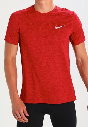 Hombre con una camiseta deportiva roja ajustada de manga corta de Nike y pantalones negros, de pie frente a un fondo liso.