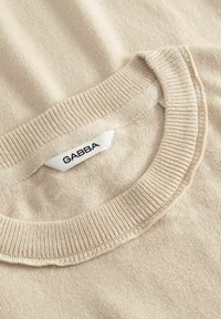 Pull en maille beige avec encolure ronde côtelée et étiquette en tissu blanc affichant « GABBA » en lettres majuscules noires.