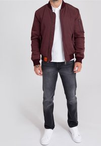 Burgundy bomberjacka med ribbad mudd och krage, som har en dragkedjeförsedd ficka på vänster ärm och en texturerad orange lappar. Bärs med grå jeans och vita sneakers.