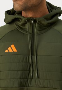 Felpa verde con zip, caratterizzata da un davanti a trapunta, tessuto texturizzato e un logo Adidas arancione sul lato sinistro. Include cordini regolabili.
