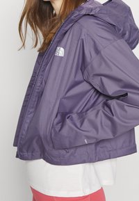 Chaqueta ligera morada con capucha, con bolsillos con cremallera y un logo en el lado superior izquierdo. Textura suave, ajuste casual.