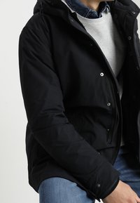 Veste matelassée noire avec capuche, dotée de fermetures à pression, d'une fermeture éclair frontale, et d'un pull gris doux en dessous. Jean bleu décontracté visible.