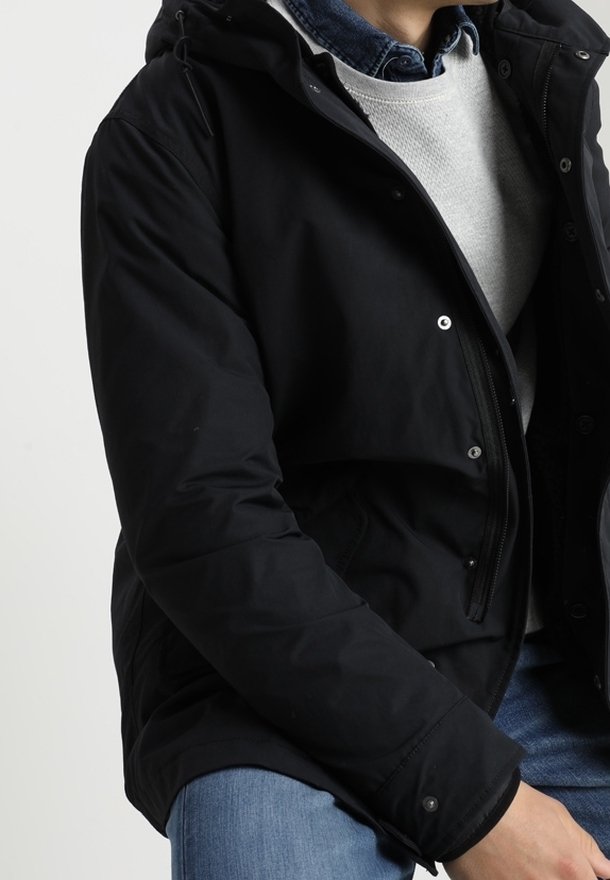 Veste matelassée noire avec capuche, dotée de fermetures à pression, d'une fermeture éclair frontale, et d'un pull gris doux en dessous. Jean bleu décontracté visible.