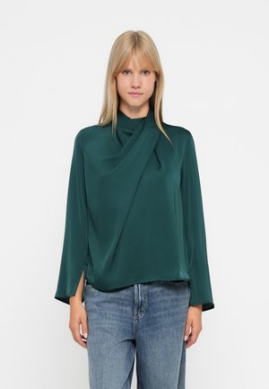 Grüner Satin-Top mit langen, ausgestellten Ärmeln, einem drapierten Ausschnitt und seitlichen Schlitzen, kombiniert mit blauen High-Waist-Jeans.