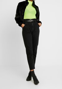 Zwarte nepbontjasje over een felgroene transparante top, hoogpols zwarte broek en enkellaarsjes. Kenmerkt zich door een ceintuur in de taille en een slim fit.