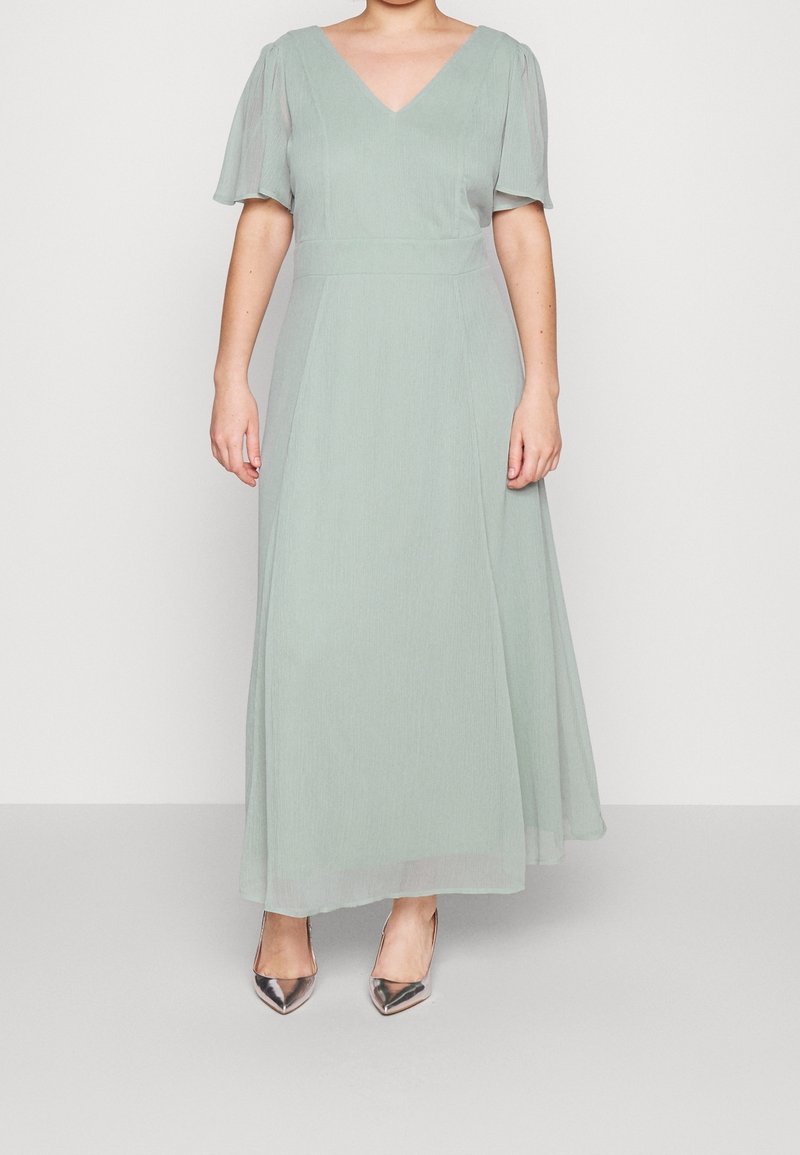 Robe longue vert clair avec un décolleté en V, des manches bouffantes courtes et une jupe fluide ; associée à des escarpins métalliques à bout pointu.