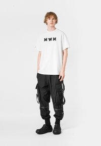 Camiseta blanca con el logo negro "MWM", combinada con pantalones cargo negros que tienen múltiples bolsillos, puños elásticos y calzado negro tipo slip-on.