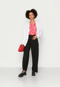 Mulher com cabelo afro, vestindo um blazer branco, uma blusa floral vermelha, calças largas pretas e sapatos pretos, segurando uma bolsa de mão vermelha.
