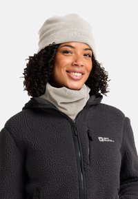Femme souriante avec des cheveux bouclés portant un bonnet en polaire gris clair et un tour de cou, ainsi qu'une veste en polaire noire zippée avec un logo.