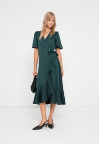 Rochie teal dintr-un material neted, cu mâneci puff scurte, un cordon lateral și o fustă cu volane în cascade. Purtați cu tocuri negre și o geantă.