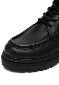 Fekete bőrcipő vastag talppal, részletes varrással és fekete fűzővel, amely fém szegecsekbe van fűzve.