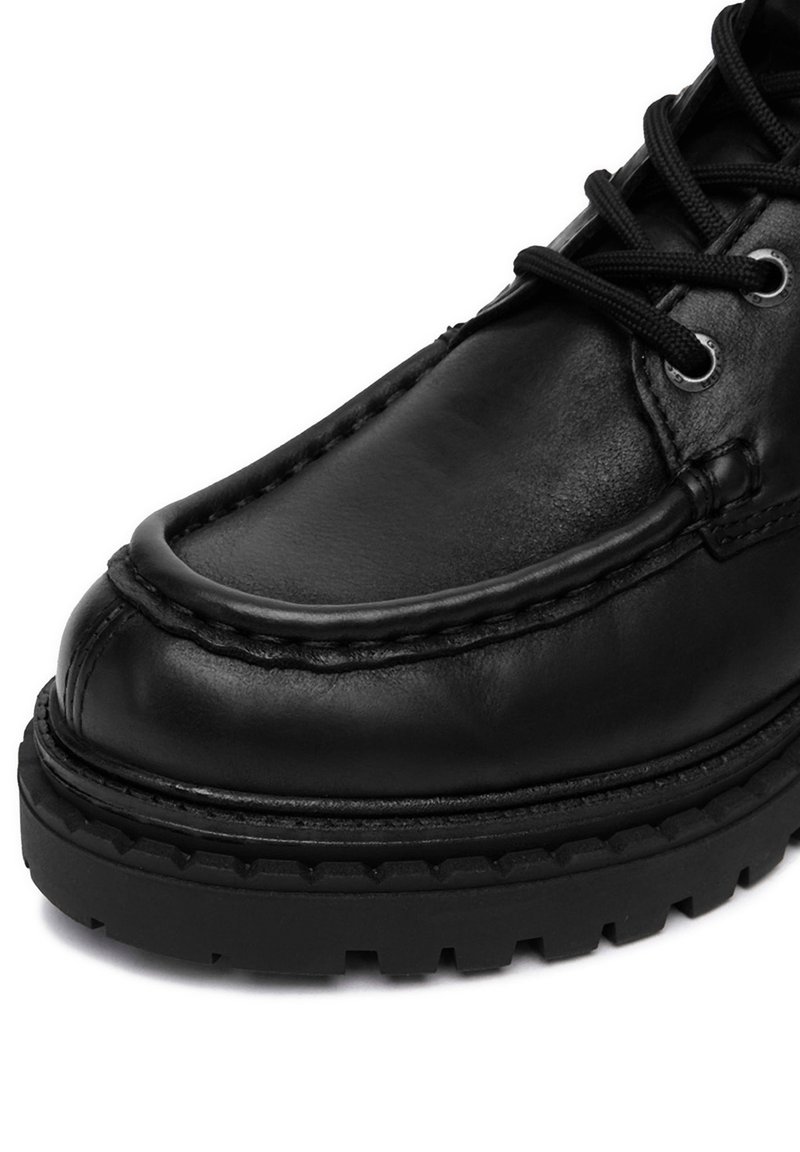 Scarpa in pelle nera con suola spessa, cuciture dettagliate e lacci neri inseriti in occhiali metallici.