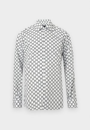 Cămașă albă cu mâneci lungi, nasturi și model geometric negru în zigzag, cu guler clasic, pe un fundal simplu gri deschis.