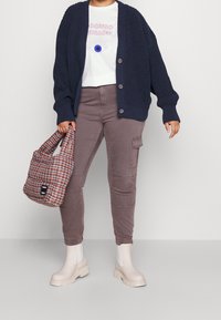 Cardigan di maglia blu navy, t-shirt bianca con grafica, pantaloni cargo grigi e stivaletti con plateau beige. Porta una borsa a tracolla a quadri con un logo.