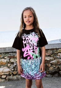 Schwarzes T-Shirt mit kurzen Ärmeln und bunten grafischen Designs, kombiniert mit einem glänzenden Paillettenrock, der von Blau zu Pink übergeht.