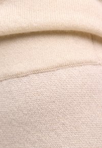 Beżowa dzianinowa tkanina swetrowa o gładkiej fakturze, z podwiniętym brzegiem i jednolitym wzorem ściegów na całej powierzchni.