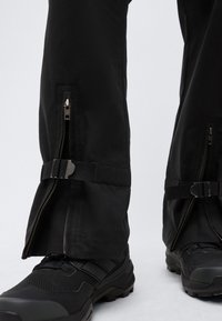 Jambes inférieures portant un pantalon noir avec des fermetures éclair latérales partiellement ouvertes au-dessus de chaussures d'extérieur noires avec des semelles texturées.
