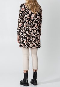 Tunique noire à motifs floraux avec des fleurs roses et beiges, manches longues, coupe légèrement oversize. Associée à un pantalon beige clair et des bottes de combat noires.