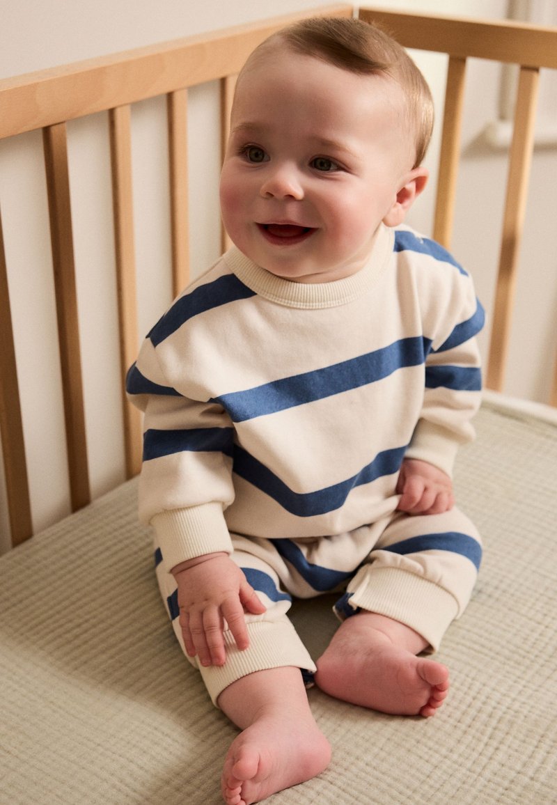 Hellbeiges Outfit mit blauen horizontalen Streifen, gerippte Bündchen, sitzt auf einer strukturierten Matratze in einem Holz-Kinderbett. Niedliches, weiches und gemütliches Design.