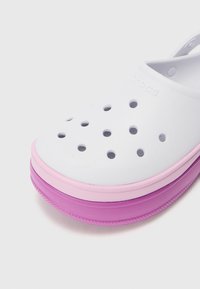 Vit plasttömningsskor med flera perforeringar, med en lagrad sula i rosa och lila. Skon har en bakrem för säker användning.