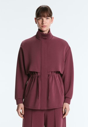 Ženska z rjavimi lasmi in šiškami, oblečena v bordo jakno z visokim ovratnikom na zadrgo z vrvico v pasu in ujemajoče hlače, stoji na belem ozadju.