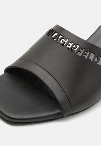 Sandale à glissière en cuir noir avec un design de logo découpé, texture lisse, bout ouvert et semelle plate de forme arrondie.