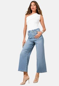 Balts vienas pleca bodijs ar pieguļošu dizainu apvienots ar gaiši ziliem augstās jostasvietas platmalu džinsiem, kuriem ir dekoratīvi elementi uz kabatas.