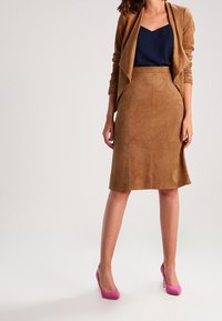 Chaqueta de ante marrón sobre una blusa azul marino, combinada con una falda de ante marrón a juego y tacones altos rosa. Textura suave con detalles de corte a medida.