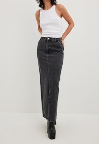 Mujer vistiendo una blusa sin mangas blanca, una falda larga de mezclilla gris oscuro de cintura alta y botas negras de plataforma, de pie contra un fondo liso.