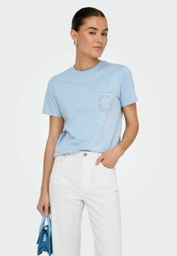 Lyseblå bomulds T-shirt med rund halsudskæring, korte ærmer og blonde-lomme detalje. Sammen med hvide højtaljede bukser og en blå håndtaske.
