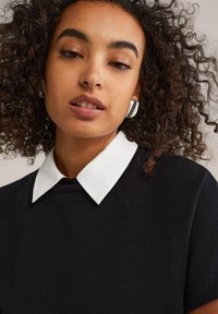 Mustassa neulepaidassa on valkoinen litteä kaulus, lyhyet hihat ja teksturoitu kangas. Mallilla on päällään hopeiset korvakorut. Taustalla on neutraali väri.
