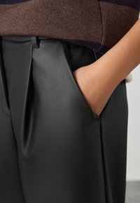 Crne hlače od umjetne kože s naboranim dizajnom i bočnim džepovima. Tekstura izgleda glatko, s krojem po mjeri i suptilnim sjajem.