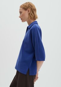Femme en haut bleu à manches trois-quarts et pantalon foncé, debout de profil sur un fond gris clair uni.