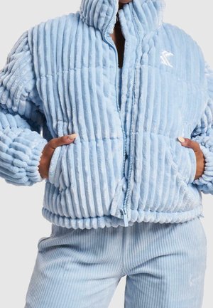 Personne portant une veste polaire côtelée bleu clair et un pantalon, avec les mains dans les poches de la veste, fermeture éclair visible et détail du col.