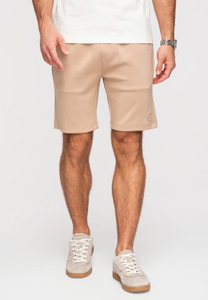 Mand iført beige shorts, hvide sneakers, hvid skjorte og sølvur står mod en ensfarvet baggrund.