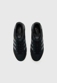 Čierne atletické topánky s vrchnou časťou zo semišu a sieťoviny. Majú tri sivé pruhy, zaoblenú špičku a tmavú gumovú podrážku. Logo na jazyku a boku.