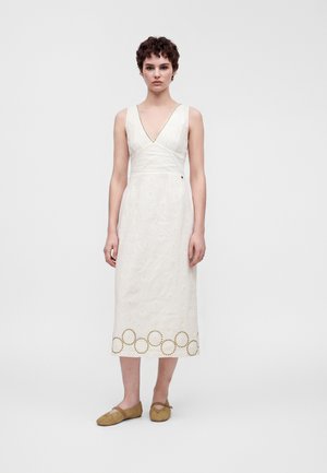 Vrouw met kort krullend haar draagt een witte mouwloze midi-jurk met een V-hals en ronde uitsnijdingen nabij de zoom, gecombineerd met beige platte schoenen.